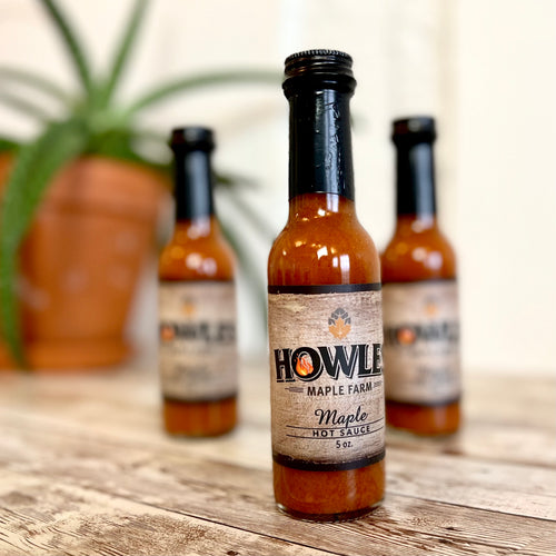 Three bottles of the howle's maple hot sauce 5 ounce bottle. Tow bottles are blurred in the background all placed on a wooden board. 