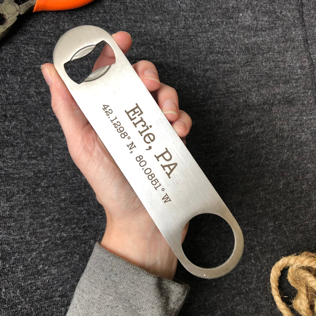 Person's hand holding silver bottle opener that reads Erie, PA and its map coordinates on a grey background 